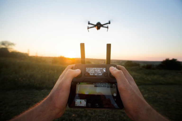 Two hands holding a remote controller with antennas and screen, flying quadcopter drone visible against a sunset sky, warm orange and gold tones